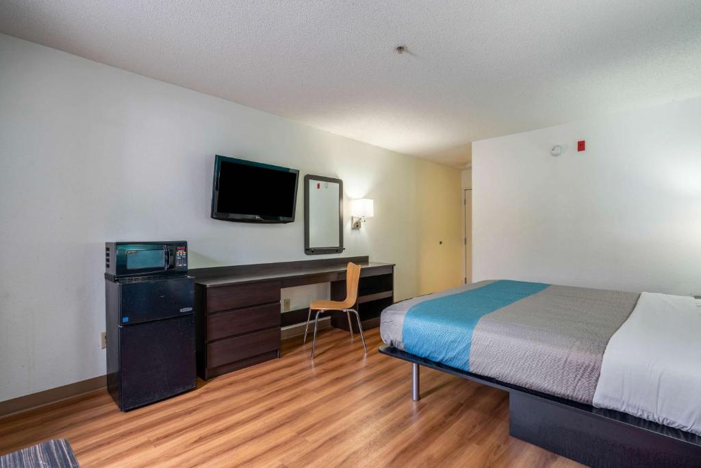 A single bed inside a room with white walls and wooden floor. A fridge, microwave oven, a TV, a desk, and a chair can be seenfacing the bed. This is in Motel 6, a pet friendly hotel in Rocky MOunt