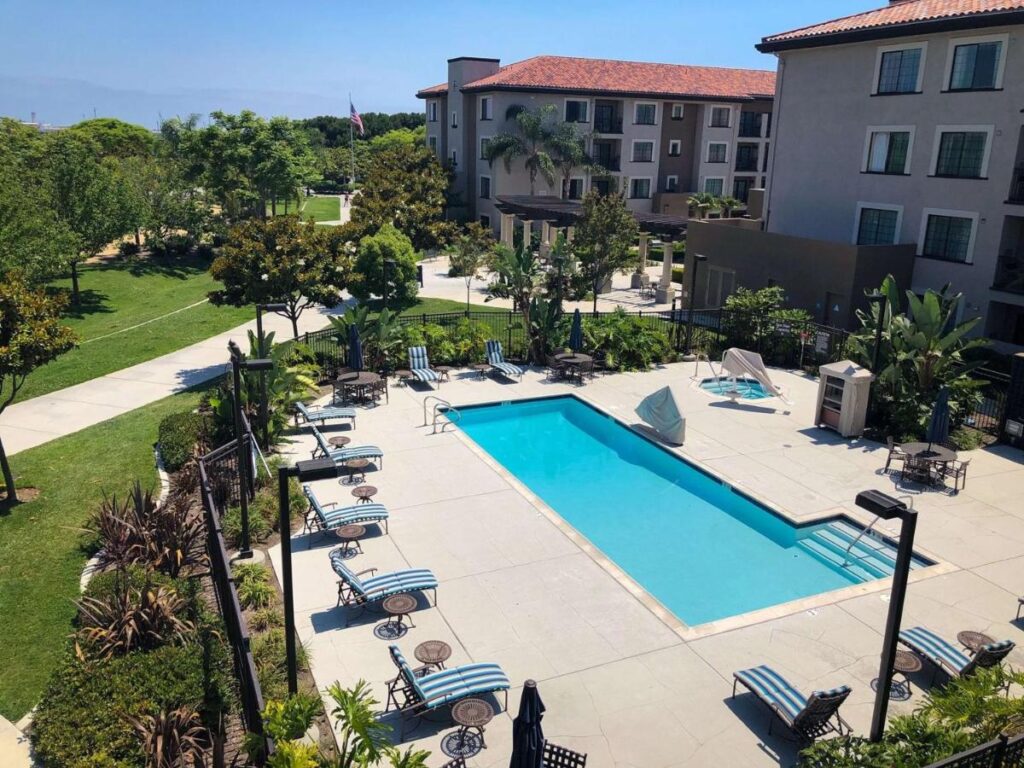 L shaped outdoor pool at Homewood Suites By Hilton San Diego with lounge chairs and tables on the side. The properrty is surrounded with different trees and grass.