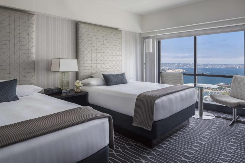 hotel room at Manchester Grand Hyatt San Diego with 2 double beds. there are 2 chairs and a table by the window. the view from the window is the harbor.
