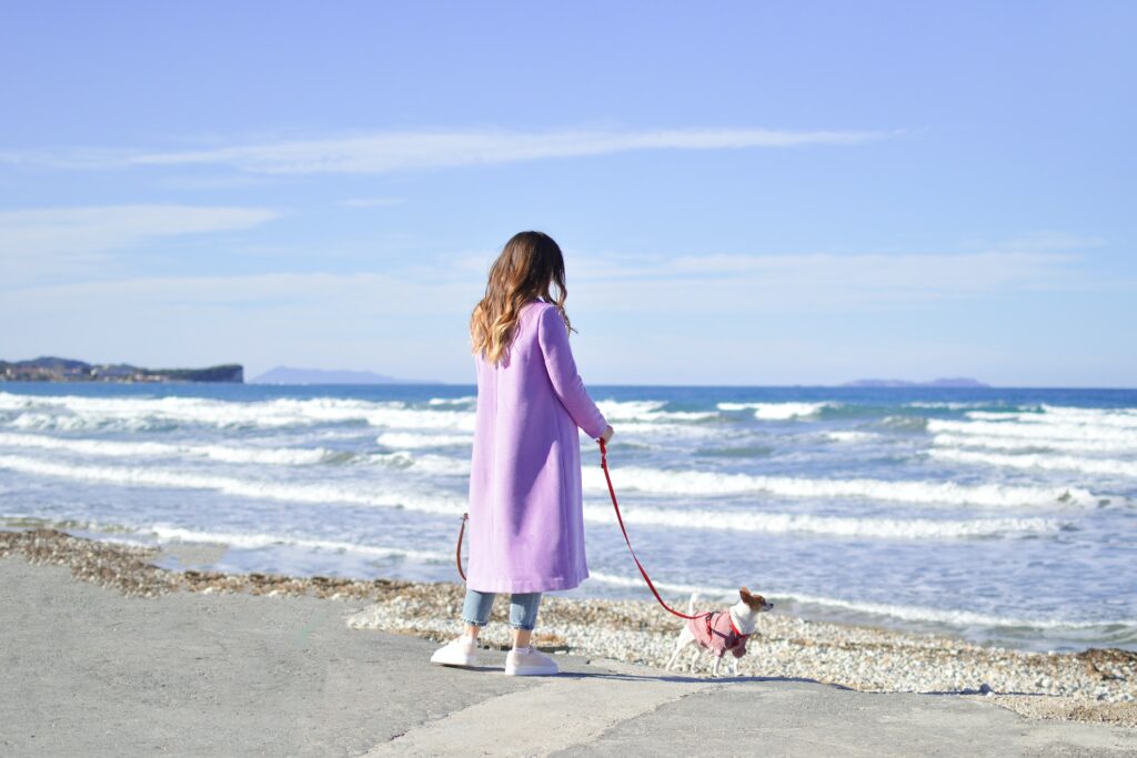 woman with brown curly hair wearing a long purple coat holding the red leash of her dog while taking a stroll at dog friendly beach in Rhode Island